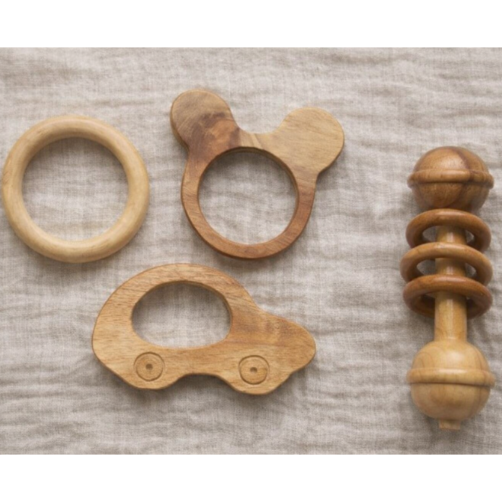 Four natural wood baby teething toys arranged on a textured beige linen fabric background. The set includes a simple ring, a teether with a looped handle, a car-shaped teether, and a spiral spool-style rattle.