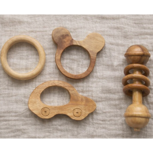 Four natural wood baby teething toys arranged on a textured beige linen fabric background. The set includes a simple ring, a teether with a looped handle, a car-shaped teether, and a spiral spool-style rattle.